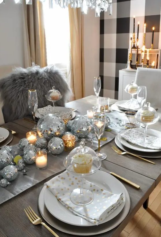 a shiny holiday tablescape with silver disco balls forming a table runner, candles, white porcelain and gold cutlery