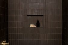 31 a refined moody bathroom clad with chocolate brown tiles, with chic gold fixtures, a white tub and a niche for storage