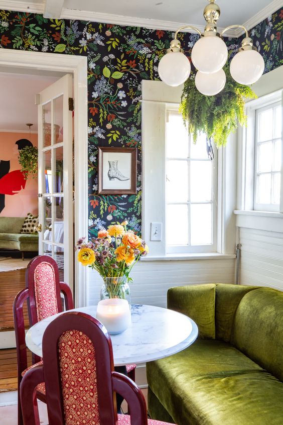 an eclectic living space with dark floral wallpaper, a chartreuse sofa, a round table, red chairs and a chandelier with greenery