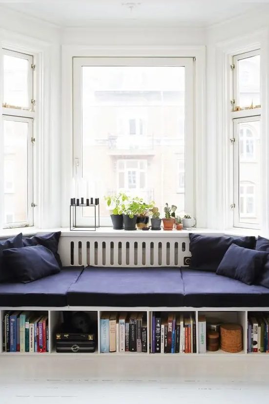 A bay window reading nook styled with midnight blue cushions and pillows, built in bookshelves, potted plants and a candelabra