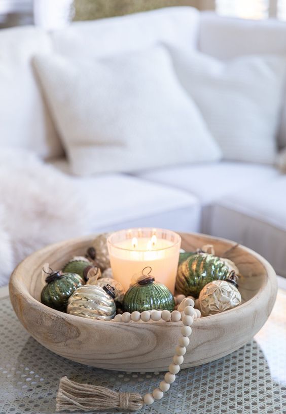 a Scandi Christmas arrangement of a wooden bowl with green and gold ornaments, a candle and wooden beads plus a tassel