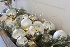 a glam Christmas arrangement of a wooden bowl with silver and white ornaments, evergreens and lights