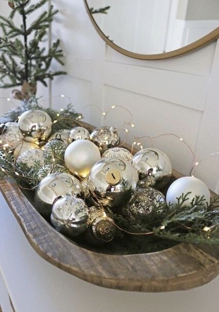 a glam Christmas arrangement of a wooden bowl with silver and white ornaments, evergreens and lights