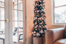a pretty slim Christmas tree with lights, white and orange ornaments and cotton branches in a basket