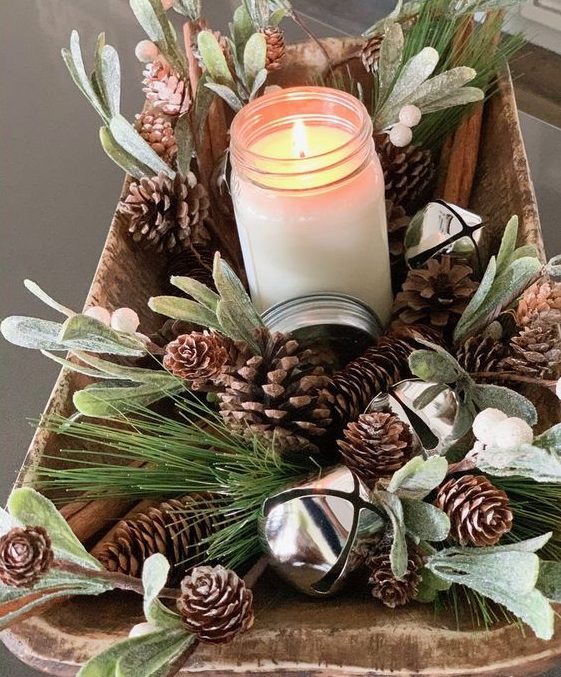 a rustic Christmas centerpiece of a wooden bowl, pinecones, greenery, bells and a candle in a jar