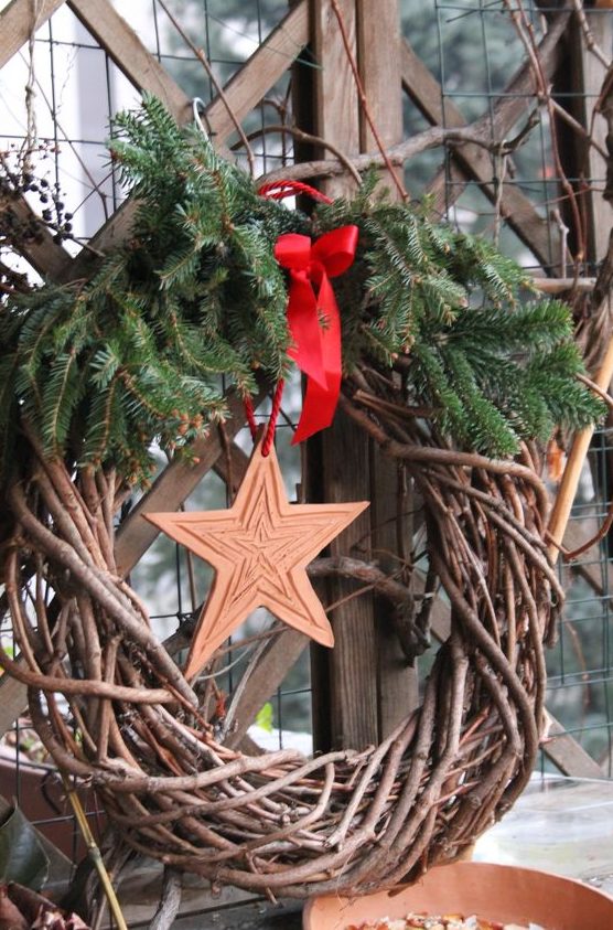 a simple rustic Christmas wreath of grapevine, evergreens, a wooden star and a red bow is a stylish and cozy idea