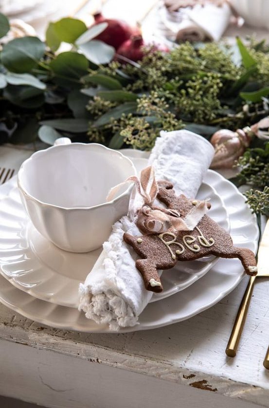 a stylish Christmas tea party table with white porcelain, a greenery and pomegranate runner, a gingerbread cookie and elegant cutlery