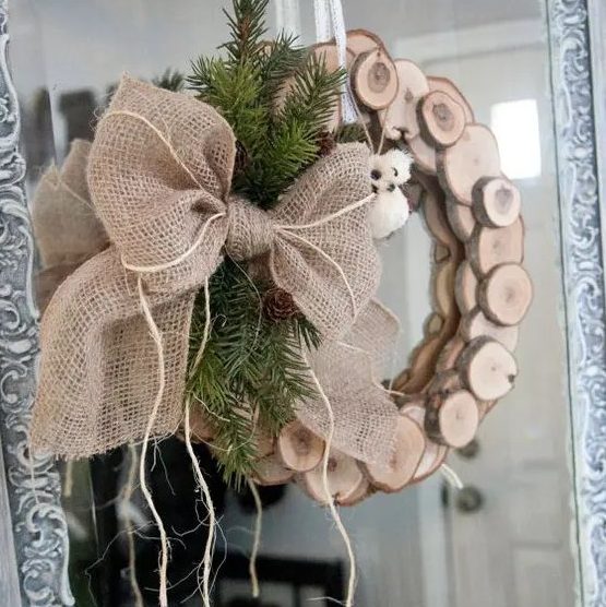 a wood slice Christmas wreath with evergreens, pinecones and a large burlap bow on the side