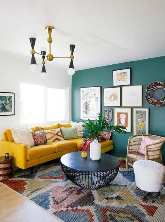 a colorful eclectic living room with a green statement wall with artworks, a boho rug, a mustard sofa and wicker touches