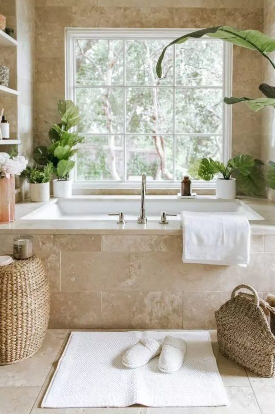a gorgeous neutral spa bathroom clad with limestone tiles, with a tub clad with them, built-in shelves and potted greenery, a woven pouf and a bag