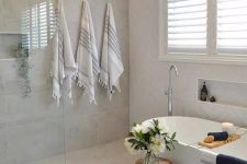 06 a modern neutral bathroom clad with neutral tiles, with an oval tub, a niche, printed and dark towels, some blooms