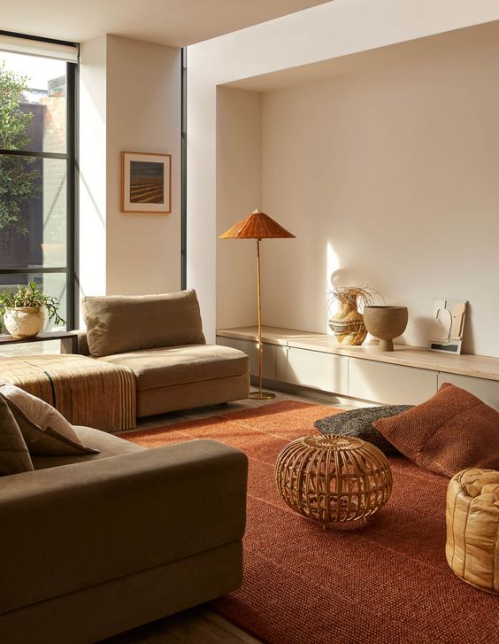 a contemporary warm earthy living room with a grey sofa and neutral seats, a deep red carpet and pillows, a floor lamp and an amber pouf