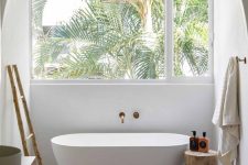 08 a neutral tropical bathroom with a window, an oval tub, a stained vanity, a ladder and a wooden stool