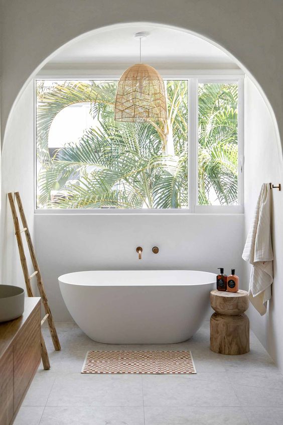 a neutral tropical bathroom with a window, an oval tub, a stained vanity, a ladder and a wooden stool
