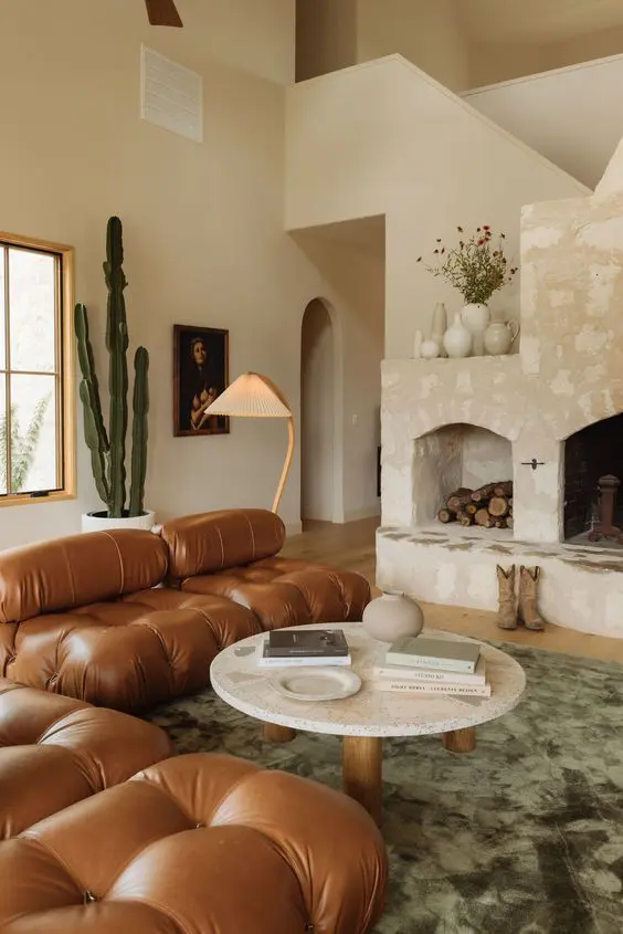 a beautiful earthy living room with a large stone hearth, a brown leather tufted sofa, a low coffee table, a green rug and plants