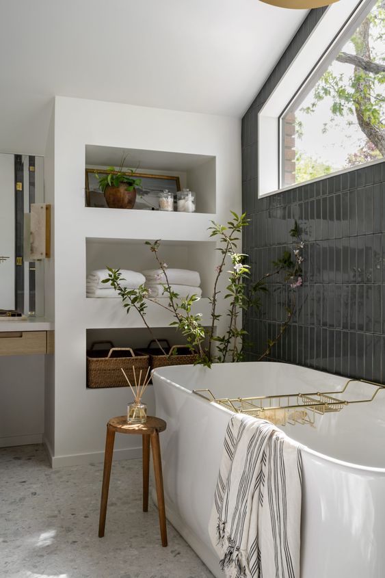A mid century modern bathroom with a window, a wall clad with black tiles, a wall with shelves, a tub and a stool