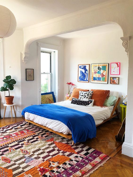 a catchy eclectic bedroom with a colorful gallery wall, a bed with bright bedding, a bold printed rug and some plants