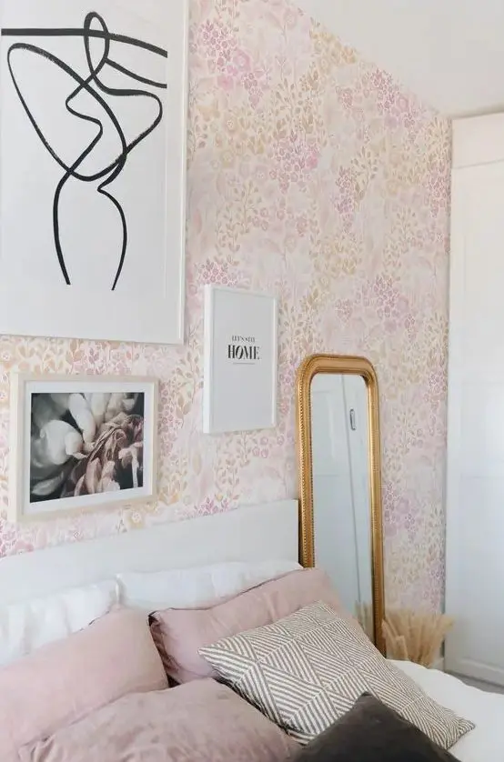 a pink bedroom with floral wallpaper, a white bed and pink and white bedding, some art and a mirror in a gold frame