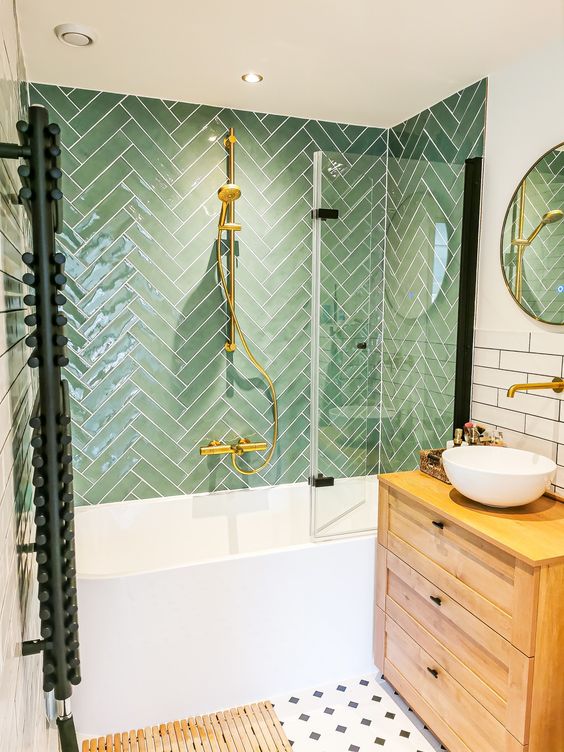 a cozy bathroom with green herringbone and white skinny tiles, a stained vanity, an oval tub and gold touches