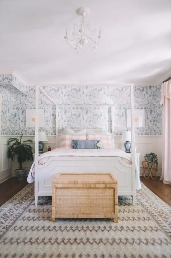 a cottage bedroom with blue floral wallpaper, a white bed with printed bedding, a woven chest, potted plants and a chandelier