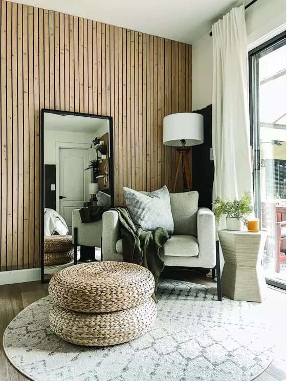 a lovely boho space with a wood slat accent wall, a neutral chair, a floor lamp, a mirror, layered rugs and jute poufs