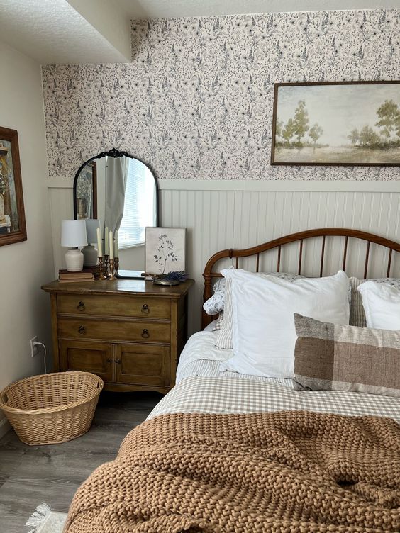 a lovely vintage cottage bedroom with floral wallpaper, paneling, a bed with printed bedding, a stained dresser, a basket and some art