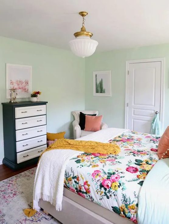 A mint colored bedroom with a bed and floral bedding, a black and white dresser, a neutral chair and a chandelier