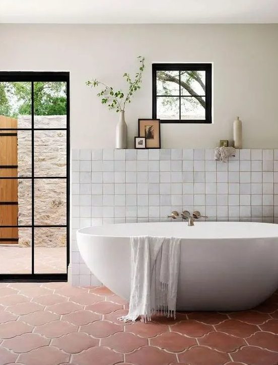 a farmhouse bathroom with neutral tiles, a terracotta tile floor, an oval tub, a glass door to the garden and neutral textiles
