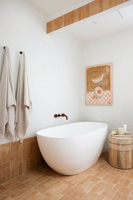 a whimsical bathroom with white and terracotta tiles, an oval tub, a side table, a bold artwork