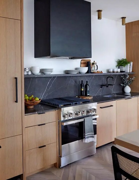 A light stained kitchen with flat panel cabinets, black soapstone countertops and a backsplash, a black hood and black hardware