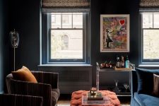 31 moody living room with navy walls, a navy sofa, a red tufted ottoman, brown chairs, some art and a bar cart