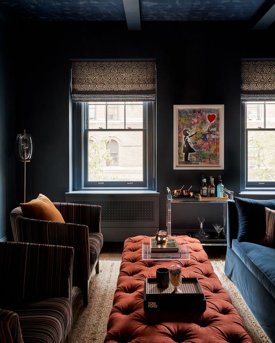 moody living room with navy walls, a navy sofa, a red tufted ottoman, brown chairs, some art and a bar cart