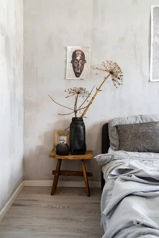 a catchy bedroom with light grey limewashed walls, a black bed with grey bedding, a wooden nightstand with dried blooms