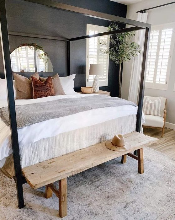 a modern farmhouse bedroom with a black accent wall, a black canopy bed, a stained bench, a potted tree and a creamy chair