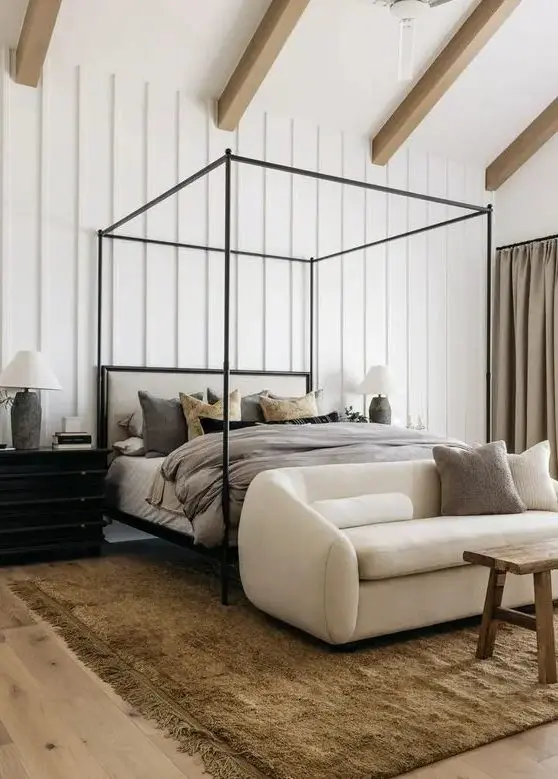 a neutral eclectic bedroom with a frame bed and grey bedding, a creamy loveseat, a stained bench, a black dresser with a lamp