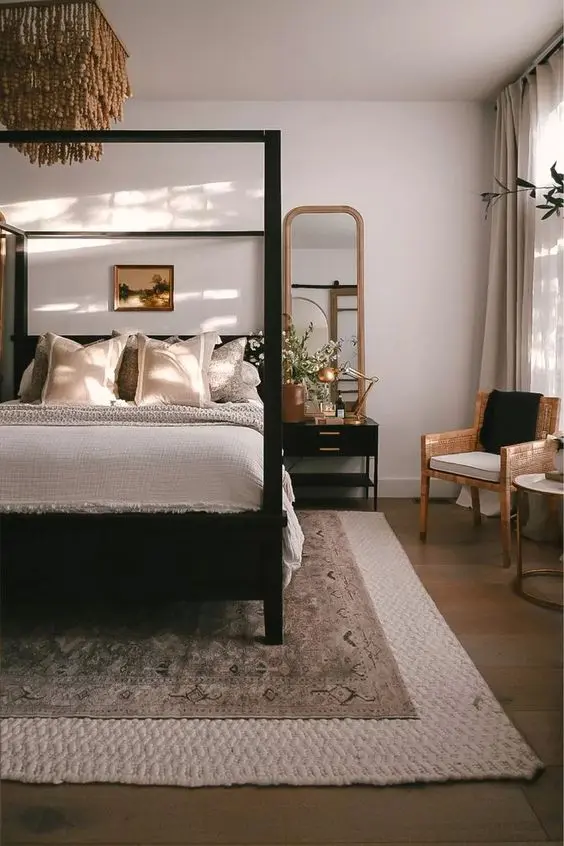 a welcoming neutral bedroom done with a black canopy bed, a chair, a black nightstand, layered rugs and a beaded chandelier