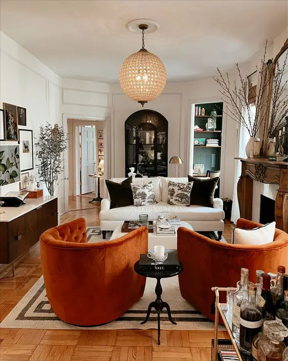 A bold living room with a fireplace and a wooden mantel, a white sofa, orange chairs, built in shelves, a credenza and a statement phere lamp