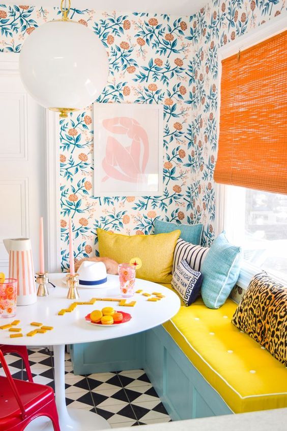 a bright breakfast book with bold floral wallpaper, a blue and yellow bench, a table, an orange shade