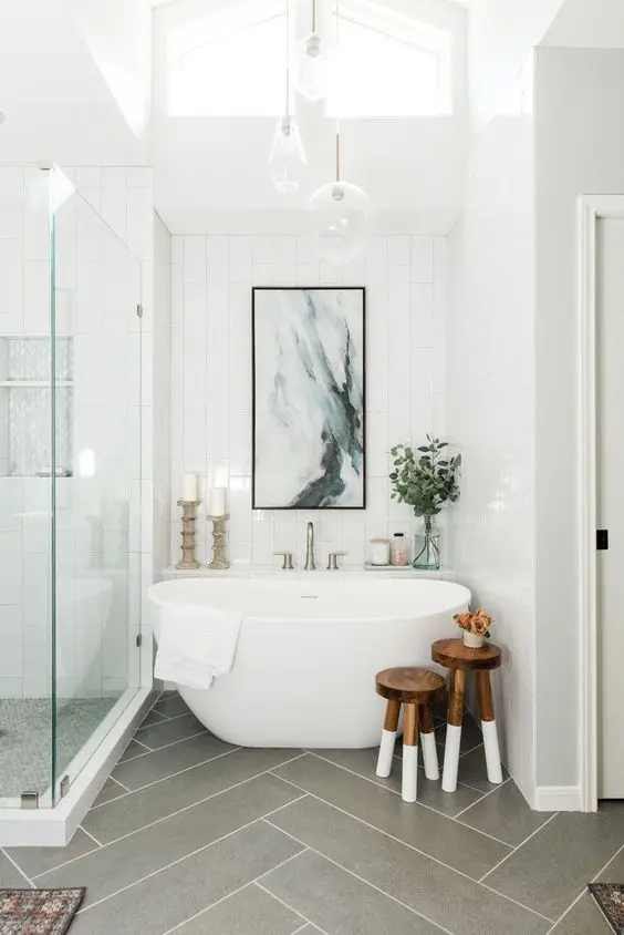 a beautiful bathroom with white stacked and grey herringbone tiles, a shower, a tub, wooden stools, an artwork and some lamps