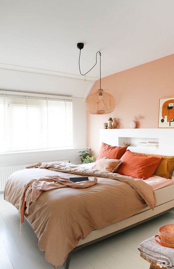 a beautiful bedroom with a peachy accent wall, a bed with a storage headboard, a woven pendant lamp and peach and coral bedding