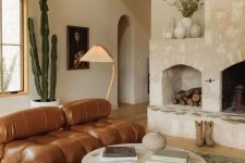 a beautiful earthy living room with a large stone hearth, a brown leather tufted sofa, a low coffee table, a green rug and plants