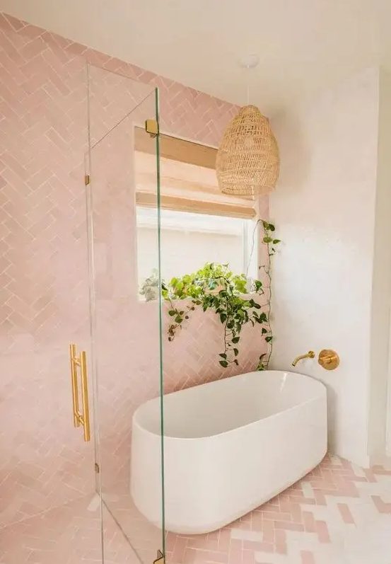 a beautiful pink and white bathroom with herringbone tiles, an oval tub, gold fixtures and a woven pendant lamp