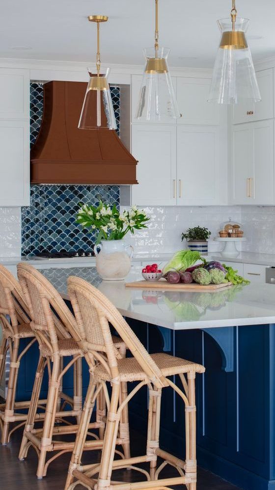 a beautiful seaside white kitchen with a navy kitchen island, a navy and blue scallop tile backsplash, a metal hood and rattan stools