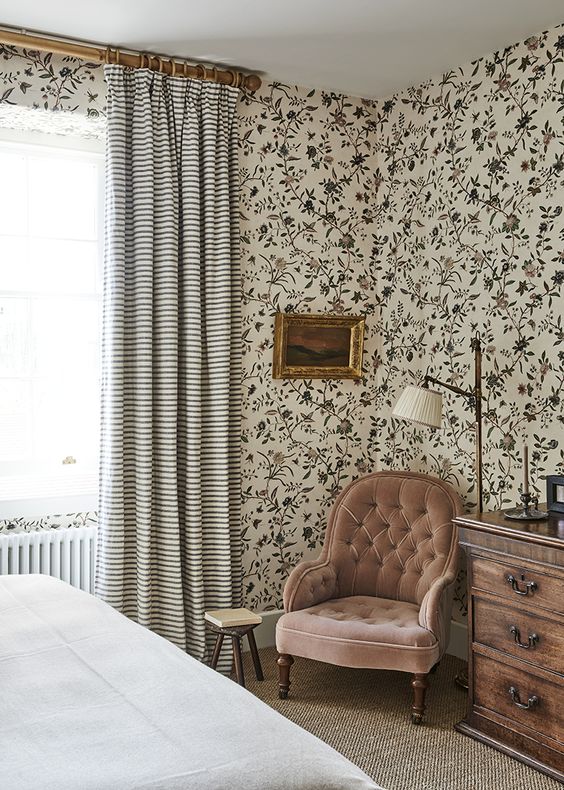 a beautiful vintage bedroom with floral wallpaper all over, a bed with neutral bedding, a blush chair, a stained dresser and a lamp