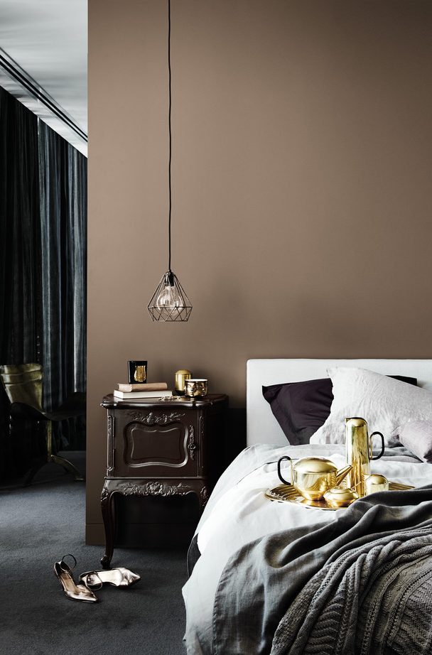 A bedroom with a brown accent wall, a bed with neutral bedding, a dark stained nightstand, a pendant lamp
