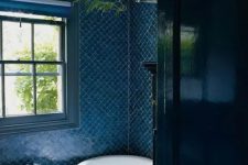 a blue fishscale tile bathroom with a free-standing tub, a black radiator and some greenery plus a window for natural light