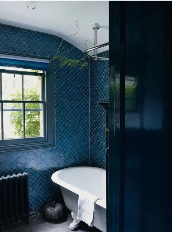A blue fishscale tile bathroom with a free standing tub, a black radiator and some greenery plus a window for natural light