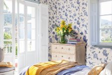 a bright and color-filled bedroom with blue floral wallpaper even on the ceiling,a bed with blue and white bedding, a shabby chic nightstand, a vintage chair