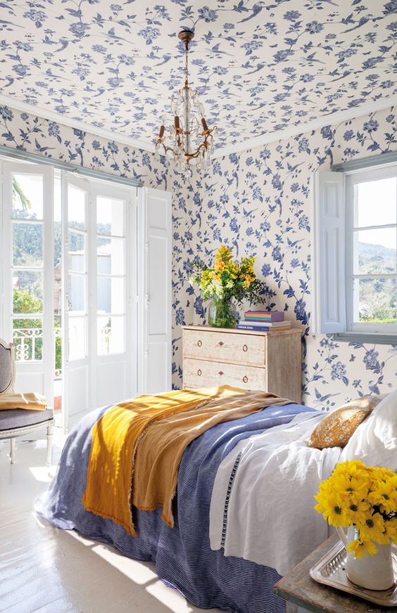 a bright and color-filled bedroom with blue floral wallpaper even on the ceiling,a bed with blue and white bedding, a shabby chic nightstand, a vintage chair