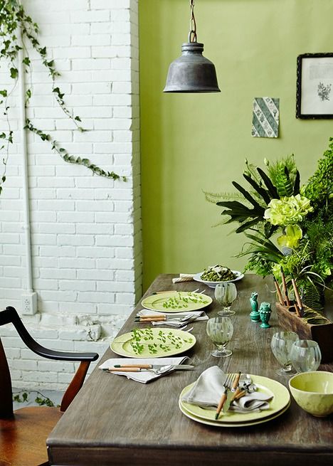 a chartreuse dining room with a brick wall, a stained table, chairs, a pendant lamp and some blooms and greenery